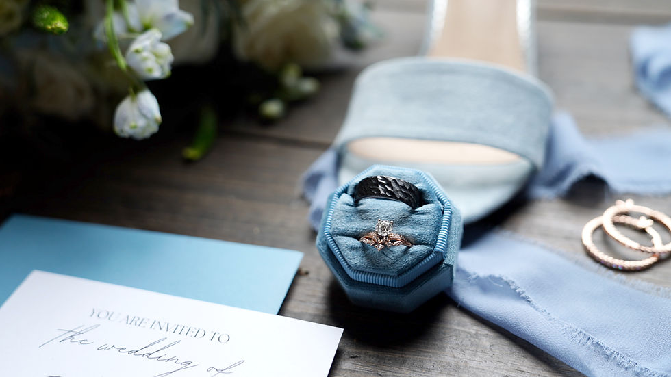 Wedding invitation with rings in a blue box, light blue shoe, and fabric on wooden table. White flowers in background. Romantic mood in Buffalo, NY.