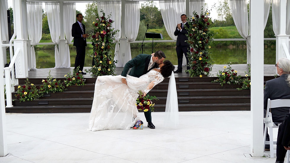 A groom dips and kisses the bride in a lacy gown, surrounded by flowers. Guests watch under a white pergola at The Ballroom at Carey Lake in Rochester NY.
