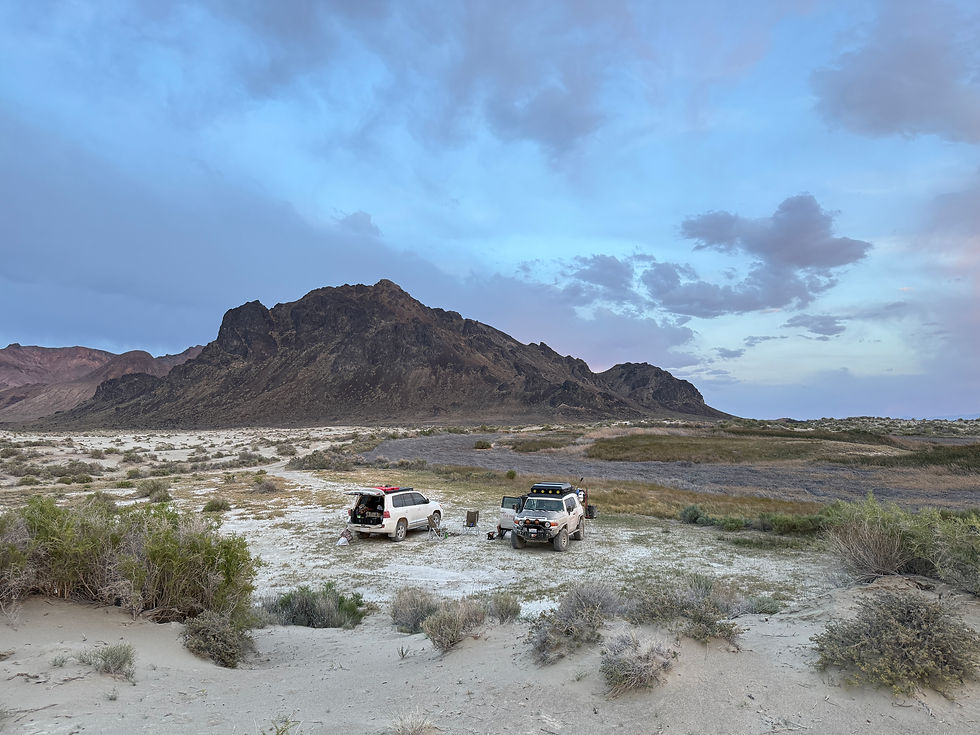 Two white off-road SUVs camp on a sandy desert plain beneath a rugged mountain and blue cloudy sky at dusk.