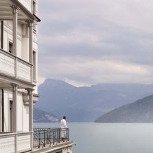 Man in a white robe standing on a balcony at Chenot Palace Weggis, gazing at the serene lake and distant mountains.