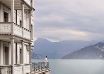 Man in a white robe standing on a balcony at Chenot Palace Weggis, gazing at the serene lake and distant mountains.
