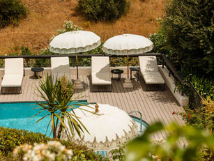 Shanti Som Wellbeing Retreat’s serene poolside deck with white sun loungers and elegant parasols, surrounded by Mediterranean greenery and dry hillside.