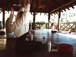 People in a wooden pavilion practice yoga, seated with arms raised. Bright light filters through trees outside. Red cushions and water bottles nearby.