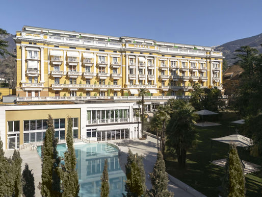 Palace Merano hotel with white balconies, palm trees, and a pool in the foreground. Mountain backdrop under a clear blue sky.