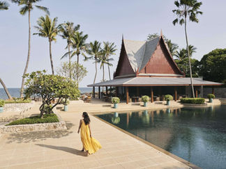 A woman in a flowing yellow dress walks along the serene poolside of Chiva-Som Health Resort in Thailand, surrounded by tropical greenery and ocean views.