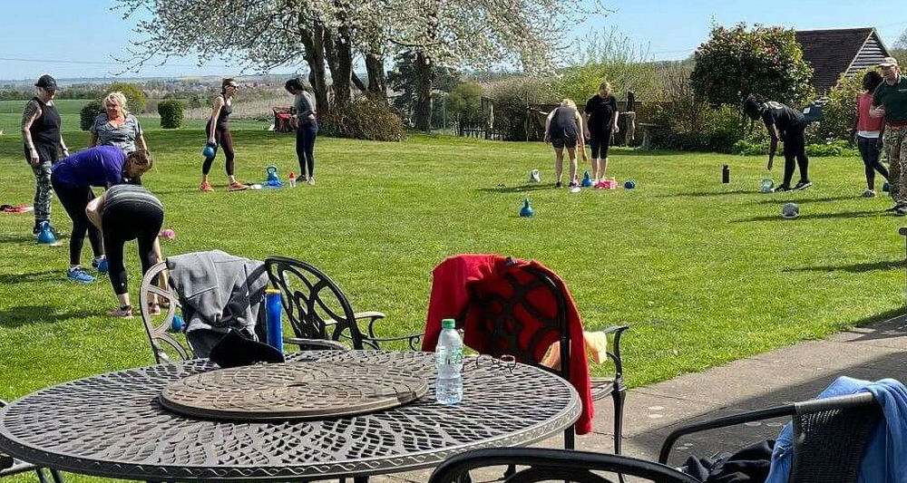 Outdoor circuit training session at GI Jane Bootcamp in England, with participants exercising on the lawn under a blossoming tree.
