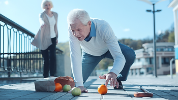Elderly man on all fours reaching for oranges on pavement. Woman behind extending hand. Dropped cane and groceries. Outdoor, sunny day.