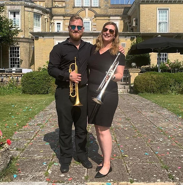 Photo of Andy and Louise dressed in black holding trumpets. They are smiling and wearing sunglasses