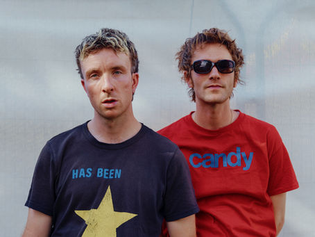 Two men pose against a pale wall, one in a navy HAS BEEN star shirt, the other in a red candy shirt and sunglasses.