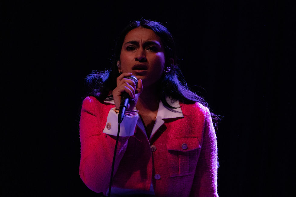 Janani K. Jha sings into a microphone on a dark stage, lit in purple and pink, wearing a bright pink jacket, focused expression.