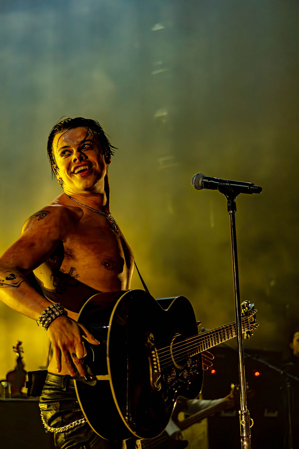 YUNGBLUD, shirtless and smiling, holds a black guitar on stage. A microphone stand is nearby. The background is illuminated in yellow.
