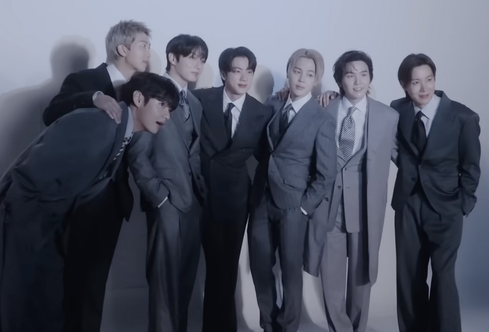 Seven members of BTS in gray suits pose together, smiling in a brightly lit studio. They're standing closely with a joyful, relaxed demeanor.
