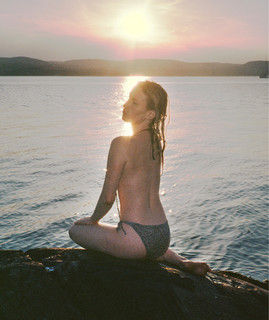 A blonde young woman at dusk in front of a body of water, topless and looking to her left