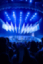Blue-lit concert stage with singer and band performing under hanging white drapes, viewed over a crowd.
