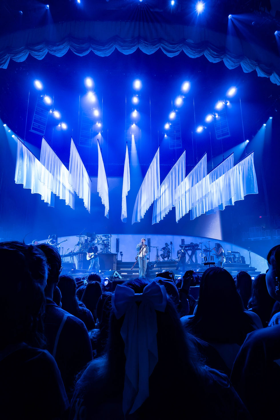 Blue-lit concert stage with singer and band performing under hanging white drapes, viewed over a crowd.