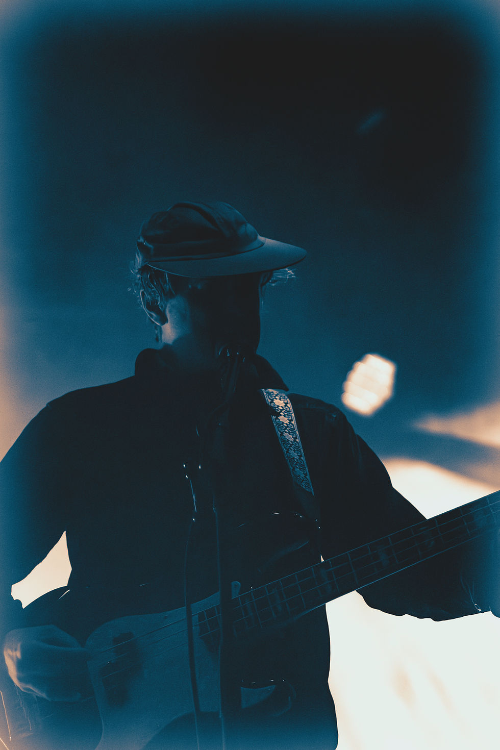Backlit musician in a cap plays guitar on a blue-lit stage, with bright lights glowing behind him.