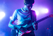 Guitarist plays an electric guitar under purple and blue stage lights, wearing a white shirt, focused and immersed in music.