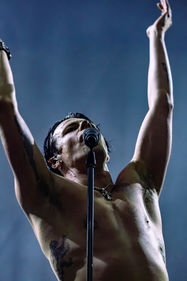 YUNGBLUD raises arms energetically, singing into a microphone on stage. Gray-blue background, vibrant live performance vibe.