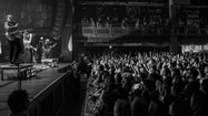 Boys Like Girls performs energetically on stage. A cheering crowd with raised hands captures the moment on phones. Dark, lively concert atmosphere.