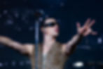 YUNGBLUD in leopard print vest and sunglasses gestures expressively on stage, holding a mic. Dark background with spotlight highlights mood.