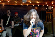 Zelda in a Led Zeppelin shirt sings into a mic, with two bandmates behind under warm string lights.