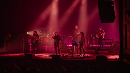 The Paper Kites performing on stage, with singer, guitars and drums under spotlights; audience silhouettes fill the foreground.