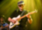 Musician wearing a cap plays an electric guitar under warm, yellow stage lights. Focused expression. Another guitarist blurred in the background.
