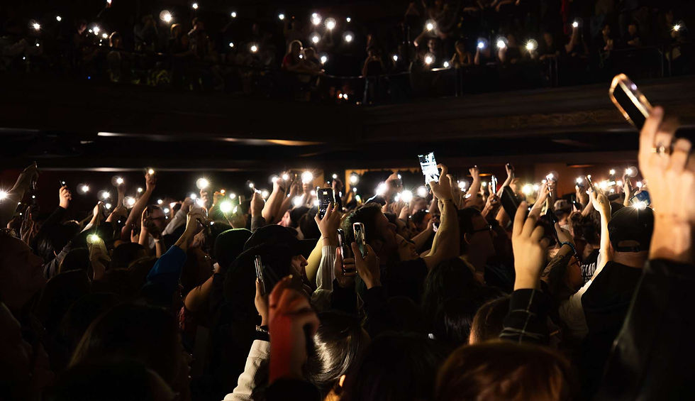 Crowd at a dark concert venue raises glowing phones, creating a sea of lights and excitement
