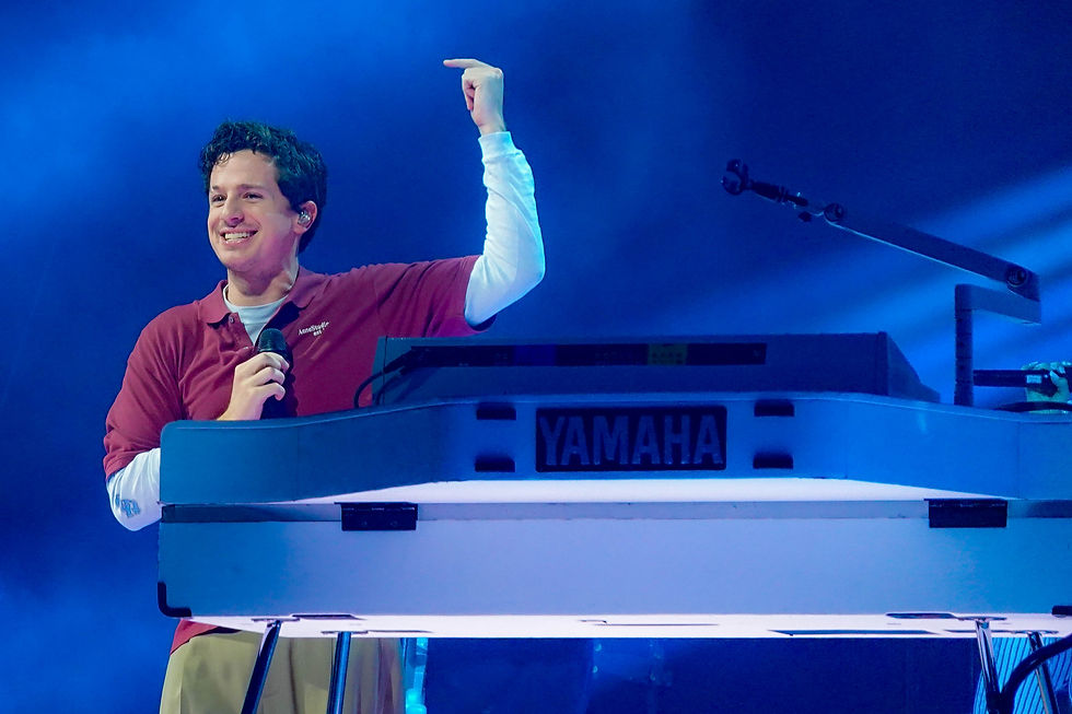 Charlie Puth points upward while holding a microphone behind a Yamaha keyboard on a blue-lit stage.
