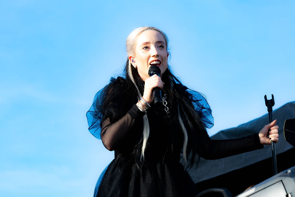 HAYLA in a black dress performs with a microphone on an outdoor stage under a bright blue sky.