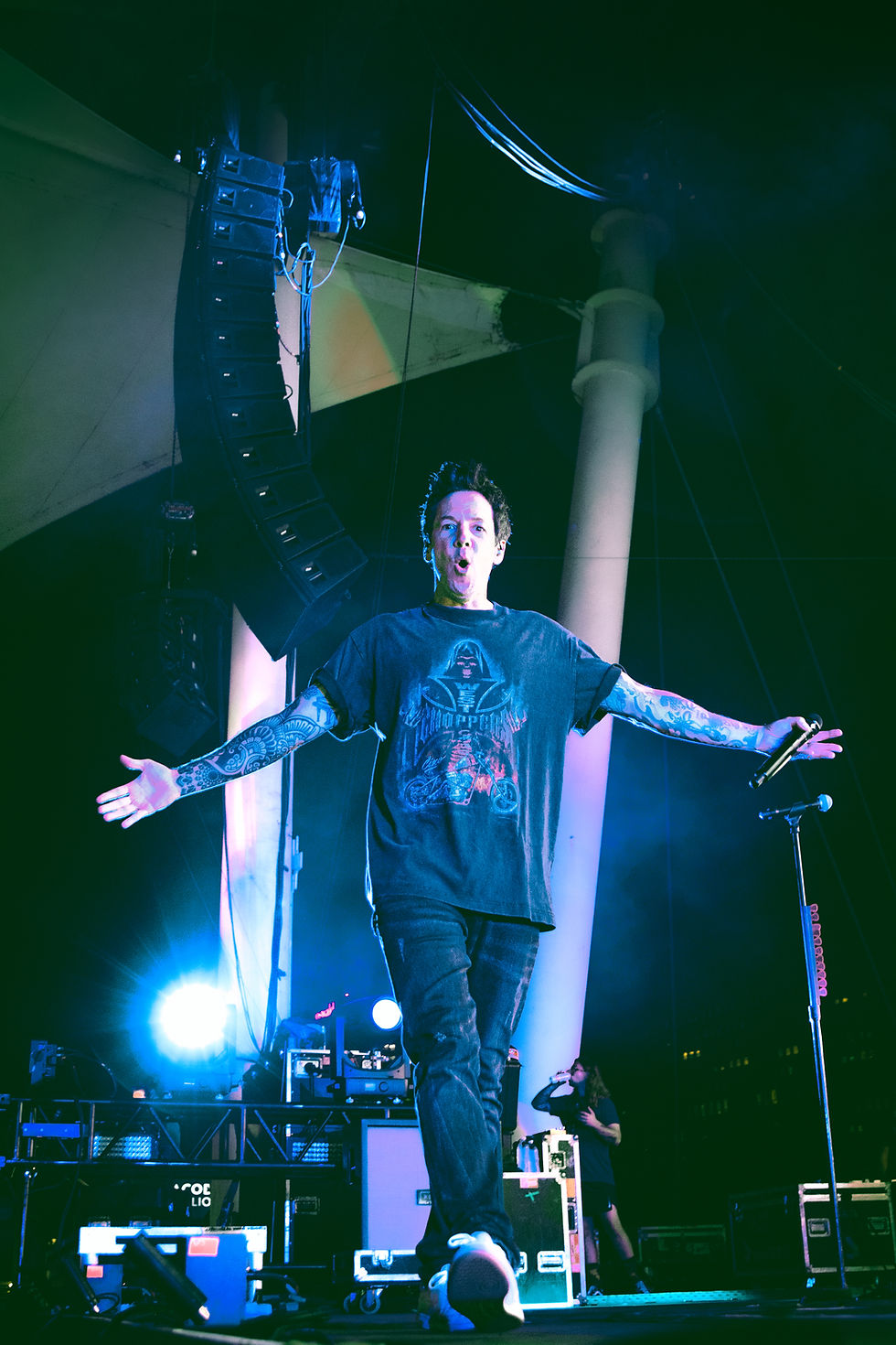 Tattooed singer strides onstage with arms out and mic in hand, under blue-green lights beside towering speakers.