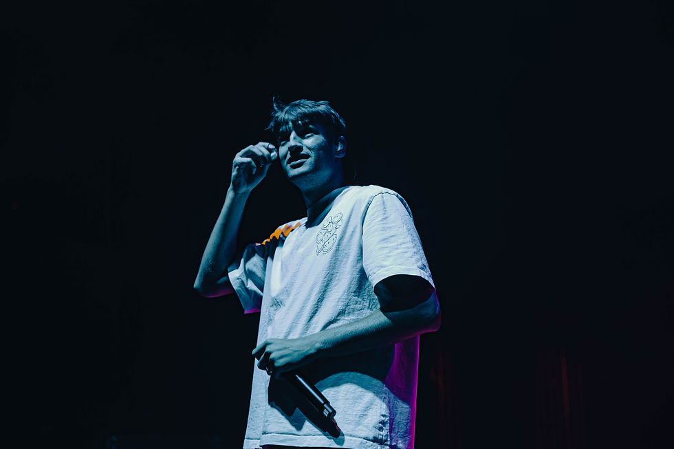 GRAHAM on a dark stage under blue and pink lights, holding a microphone and touching his face, looking focused.