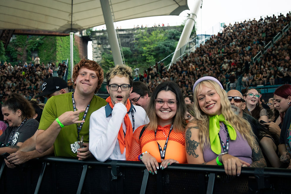 Smiling fans in colorful outfits lean on the front rail at an outdoor concert amphitheater packed with cheering crowd.
