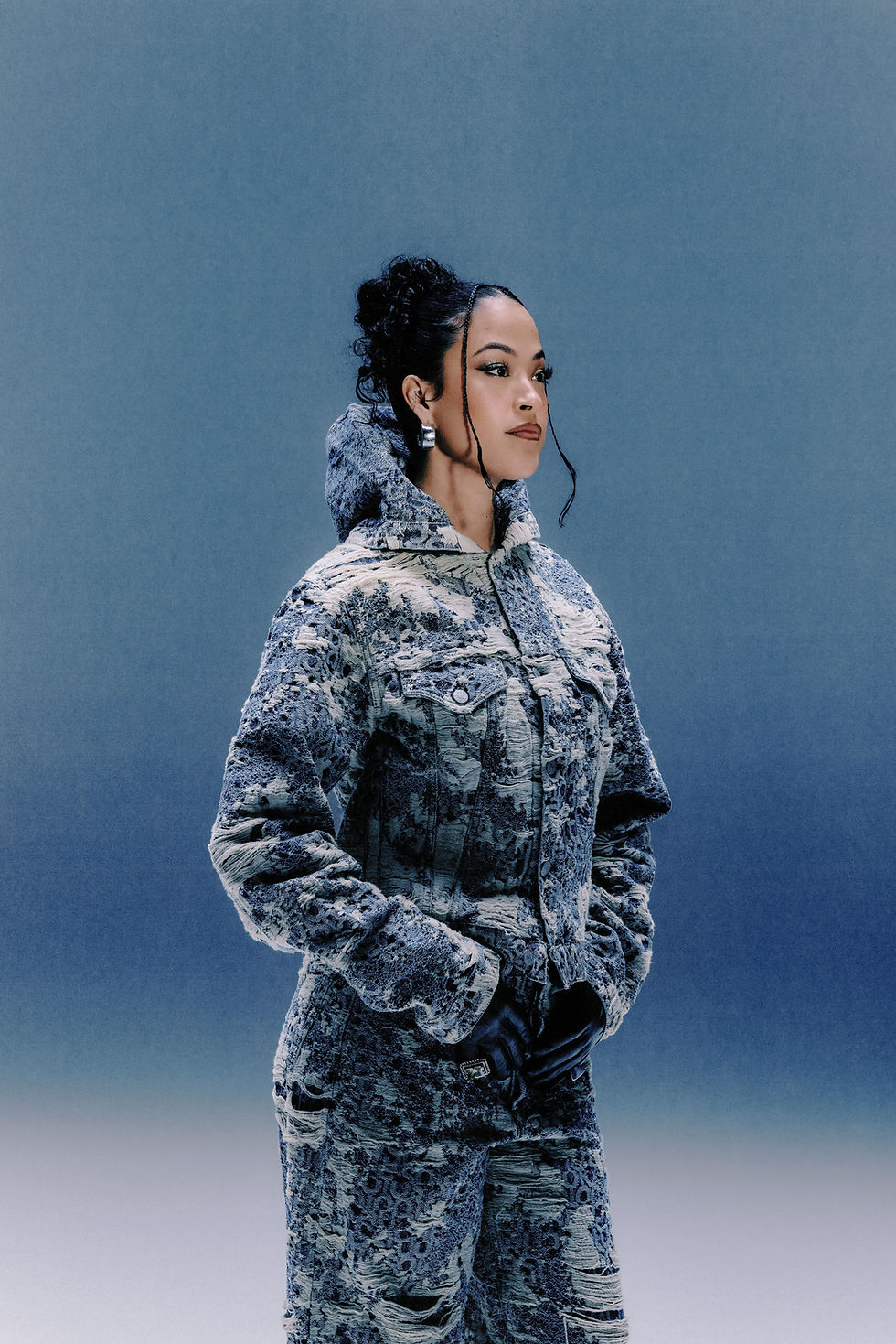 Jazzy in a textured blue patterned coat and gloves poses against a plain blue backdrop, gazing off to the side.