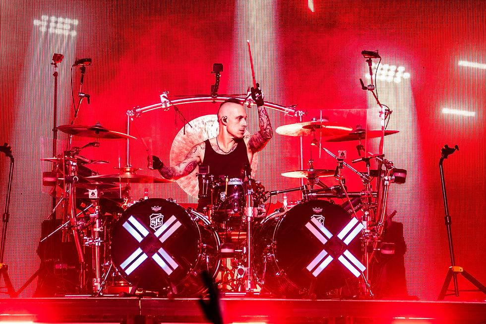 Tattooed drummer raises a stick behind a huge drum kit on a red-lit concert stage, with glowing Xs on the bass drums.