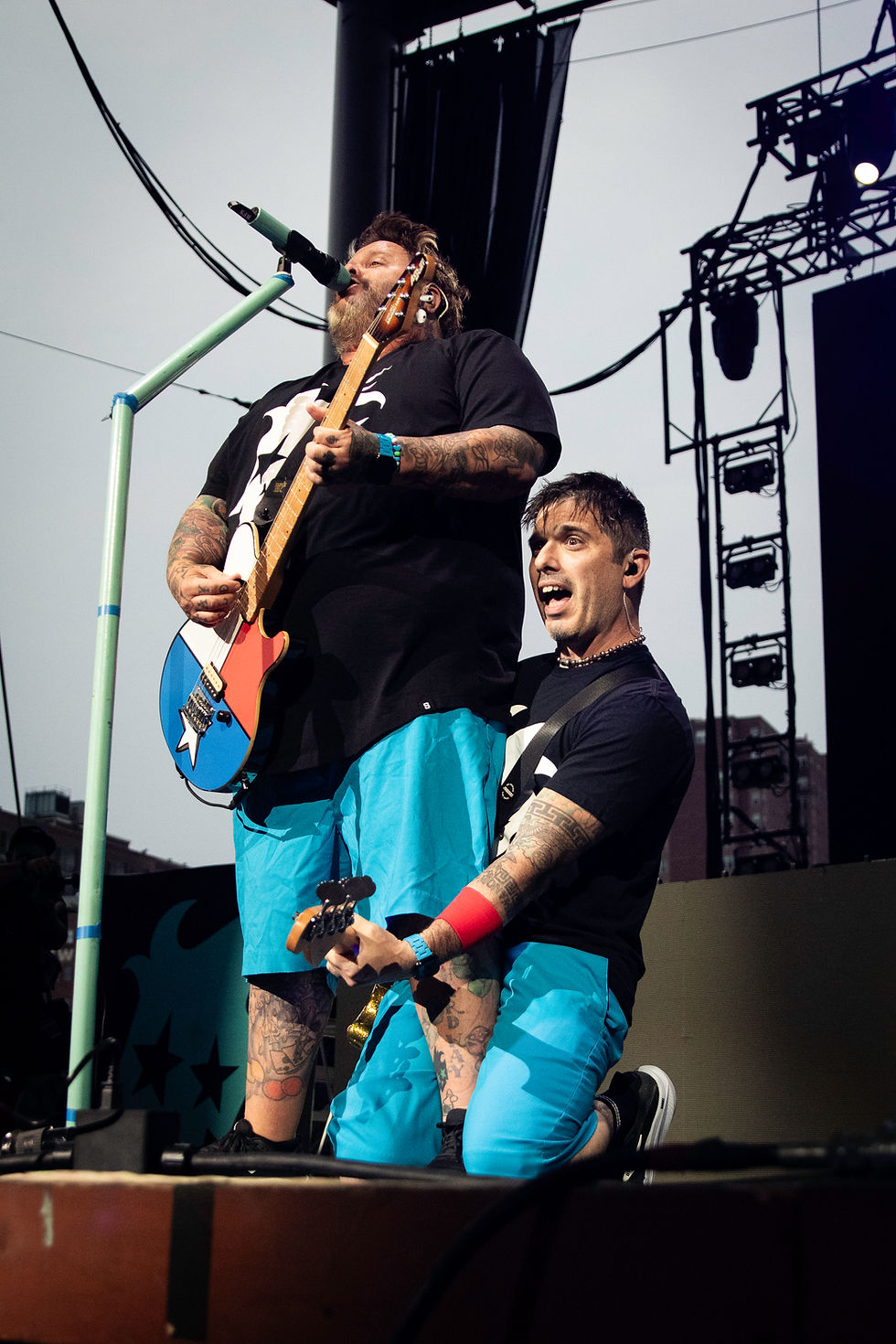 Tattooed musicians perform on an outdoor stage, one singing with a colorful guitar while another kneels beside him.