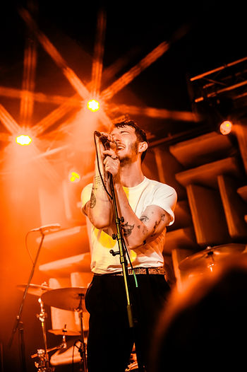 Franklin Jonas belts into a mic on an orange-lit stage, with drums behind him and bright starburst spotlights.