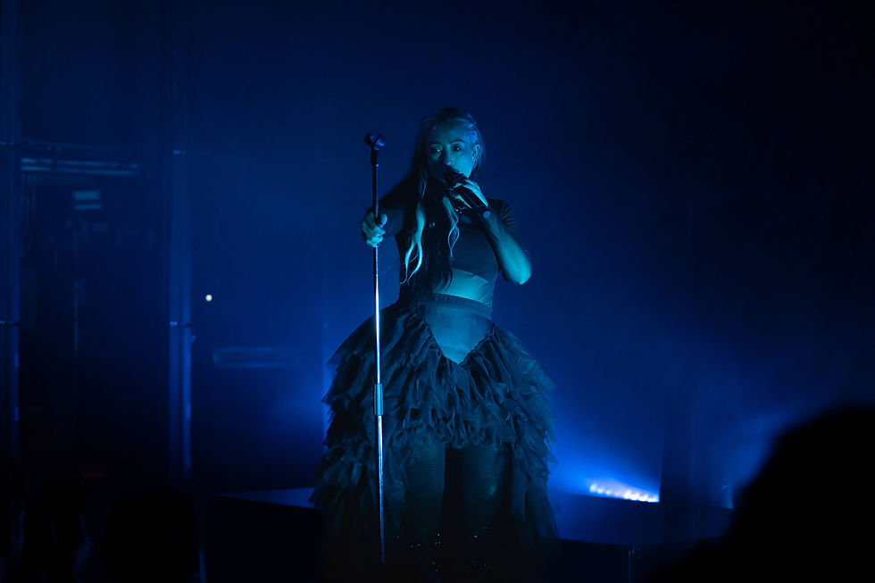 Blue-lit singer in a black ruffled dress holds a microphone onstage, singing into the crowd.
