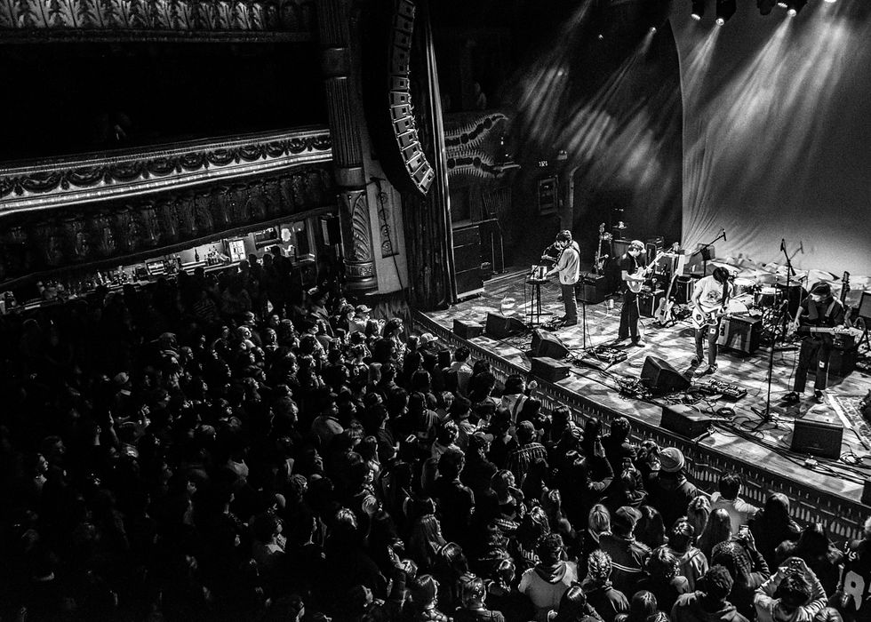 A band performs on stage in a grand theater with ornate decor. A large audience watches under dramatic stage lighting.