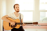 Blake Mills playing guitar in a bright room with large windows, wearing a white t-shirt, sitting in front of a pink tiled wall.