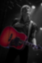 Martin Johnson with a red acoustic guitar performs on stage, hand on chest, wearing a mesh shirt. Dramatic lighting creates a moody atmosphere.