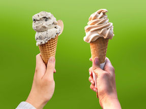 Two hands hold ice cream cones against a green background; one with cookies and cream, the other with soft-serve vanilla.