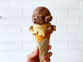 Hand holding an ice cream cone with chocolate and coffee scoops against a white brick wall. The cone is wrapped in a paper napkin.