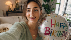 Woman recording a cheerful birthday video message selfie while holding a Happy Birthday balloon in a bright living room.