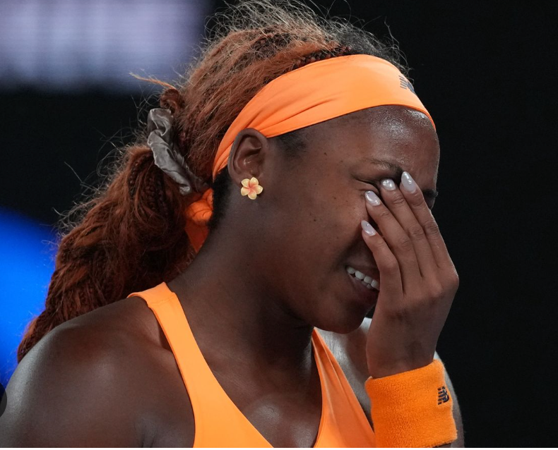 Coco Gauff, a Black woman tennis player wearing an orange headband and matching athletic outfit, covers part of her face with her hand while smiling slightly. She appears emotional during a moment on the court, with stadium lighting visible in the background.