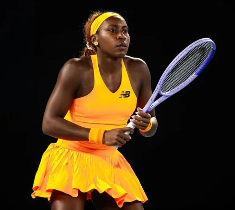 Coco Gauff, a Black woman tennis player, stands in an athletic ready position holding a tennis racket. She wears a bright orange and yellow tennis dress, a matching headband, and wristbands. Her expression is focused and serious against a dark background.