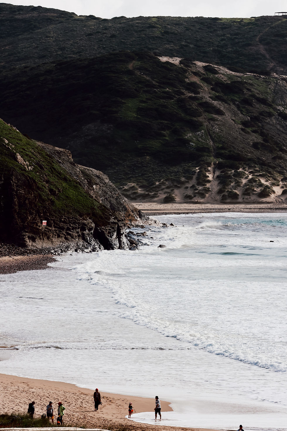 Menschen spazieren am Sandstrand nahe rauen Klippen am Meer. Im Hintergrund grüne Hügel. Die Szenerie wirkt ruhig und idyllisch.
