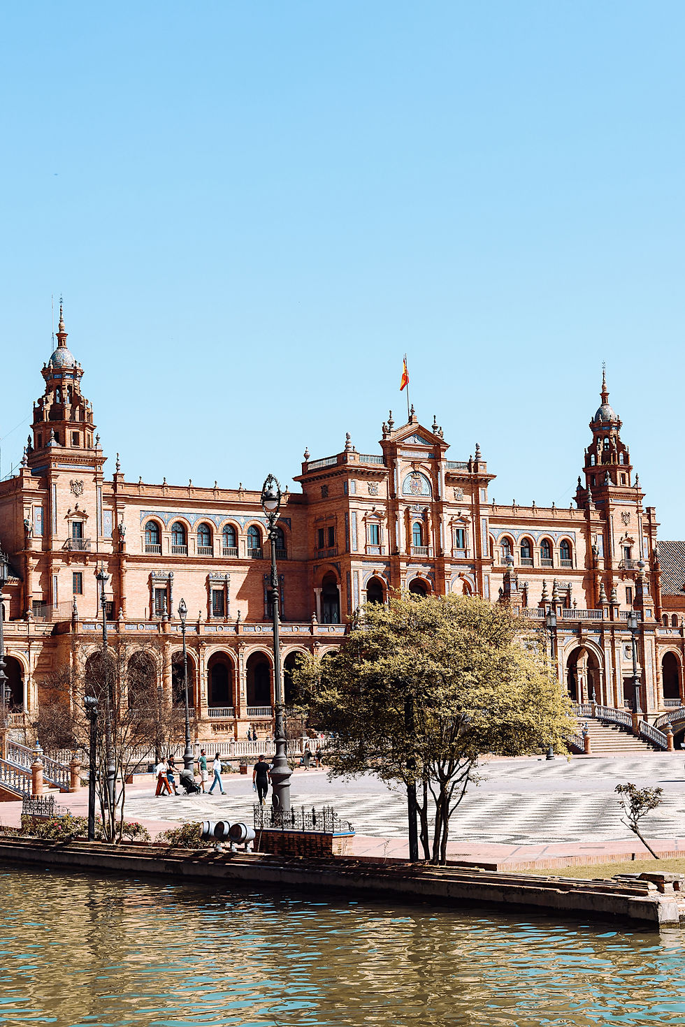 Rotes Gebäude mit Türmen im sonnigen Sevilla vor blauem Himmel. Menschen spazieren auf dem Platz, ein Kanal im Vordergrund.