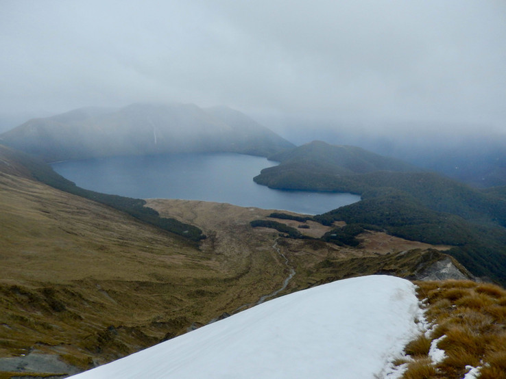 Widok z okolic Mt Burns Tarns Track na jezioro Green Lake otoczone górami i lasem, pochmurna pogoda, gdzieniegdzie śnieg, Fiordland National Park, Southland, wyspa południowa, Nowa Zelandia