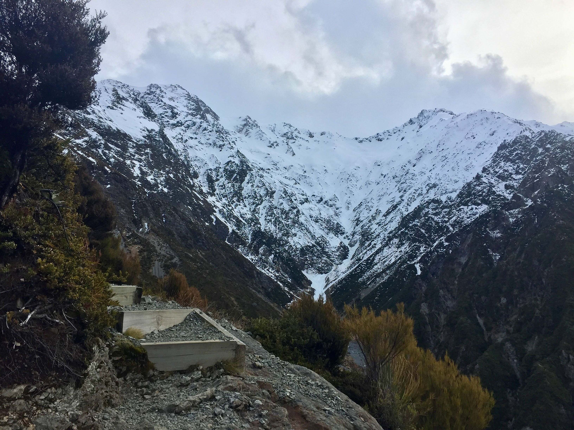 Widoki w trakcie podjeścia po szlaku Red Tarns Track w Parku Narodowym Aoraki Mt Cook w Canterbury na południowej wyspie Nowej Zelandii, w oddali pokryte śniegiem góry, żleb, na pierwszym planie drewniane stopnie na ścieżce, dookoła roślinność górska